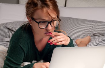Beautiful woman in a glasses with romantic hairstyle, lying on a bed with laptop, watching movie on computer
