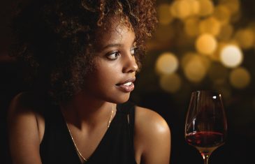 Profile of beautiful African woman with stylish curlly hair wearing elegant black dress enjoying red wine siting at restaurant having romantic dinner with husband celebrating wedding anniversary