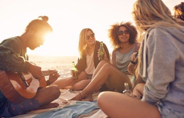 Group of young people sitting at the beach together while young man playing guitar. Group of friends partying on the beach at sunset.