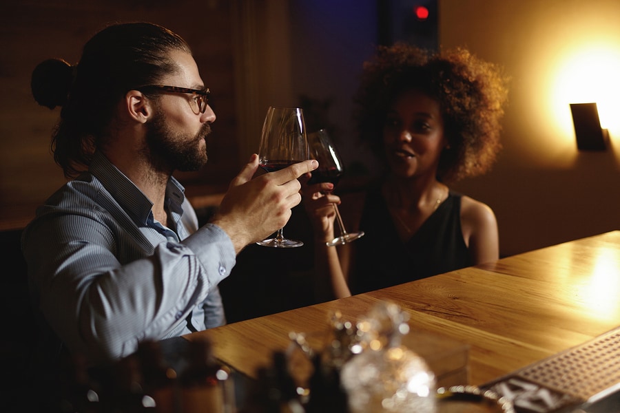 Fashionable interracial couple drinking wine during date sitting at restaurant having romantic evening and nice conversation raising glasses to love at first sight. Hipster man proposing toast